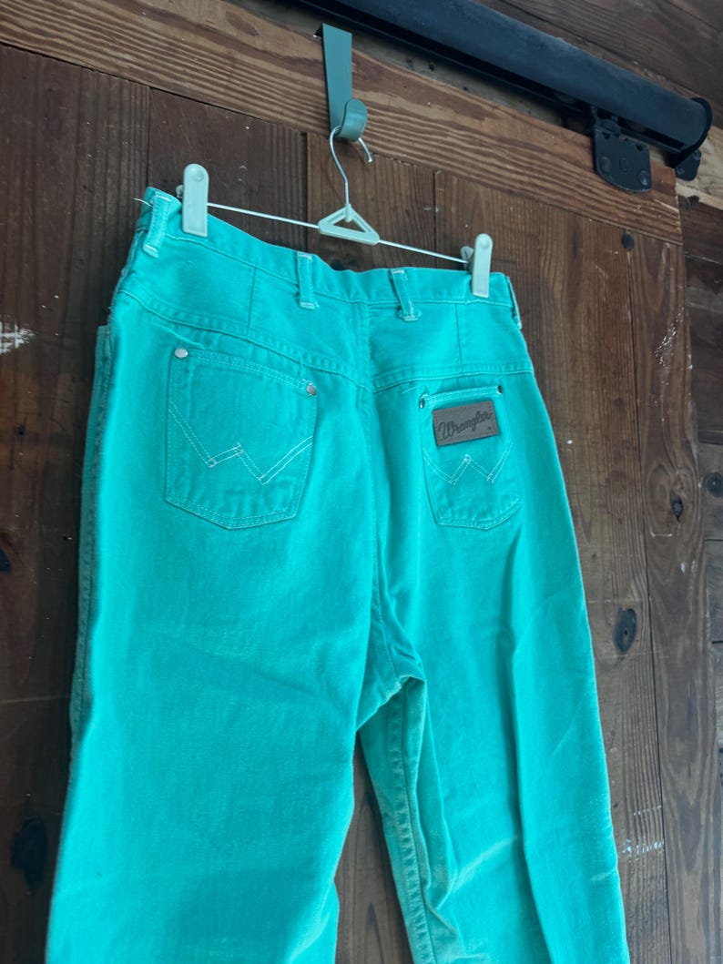 May include: Turquoise Wrangler jeans hanging on a white hanger. The jeans have a classic five-pocket design with white stitching and a brown leather patch with the Wrangler logo on the back pocket. The jeans are made of a sturdy denim material.