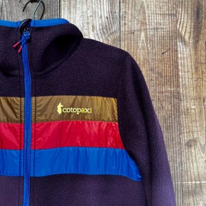 Puede incluir: Una chaqueta de forro polar con capucha en color morado oscuro que cuenta con una cremallera azul brillante. El pecho presenta franjas horizontales de colores bronce, rojo y azul, con el texto amarillo "cotopaxi" y un logo de llama impreso.