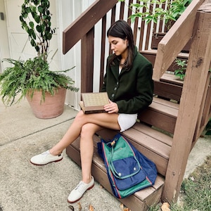 May include: A dark green blazer with three buttons, paired with white shorts and white shoes with red soles. A blue and green backpack with red trim sits on the wooden steps. A book is held in the lap.