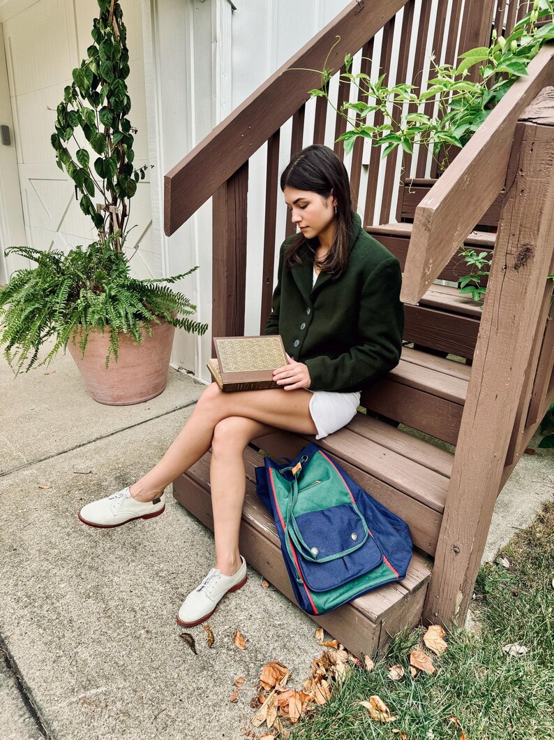 May include: A dark green blazer with three buttons, paired with white shorts and white shoes with red soles. A blue and green backpack with red trim sits on the wooden steps. A book is held in the lap.