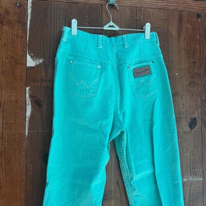 May include: Turquoise Wrangler jeans hanging on a metal hanger. The jeans have two back pockets with the Wrangler logo on the right pocket. The jeans are made of a sturdy material and have a classic fit.