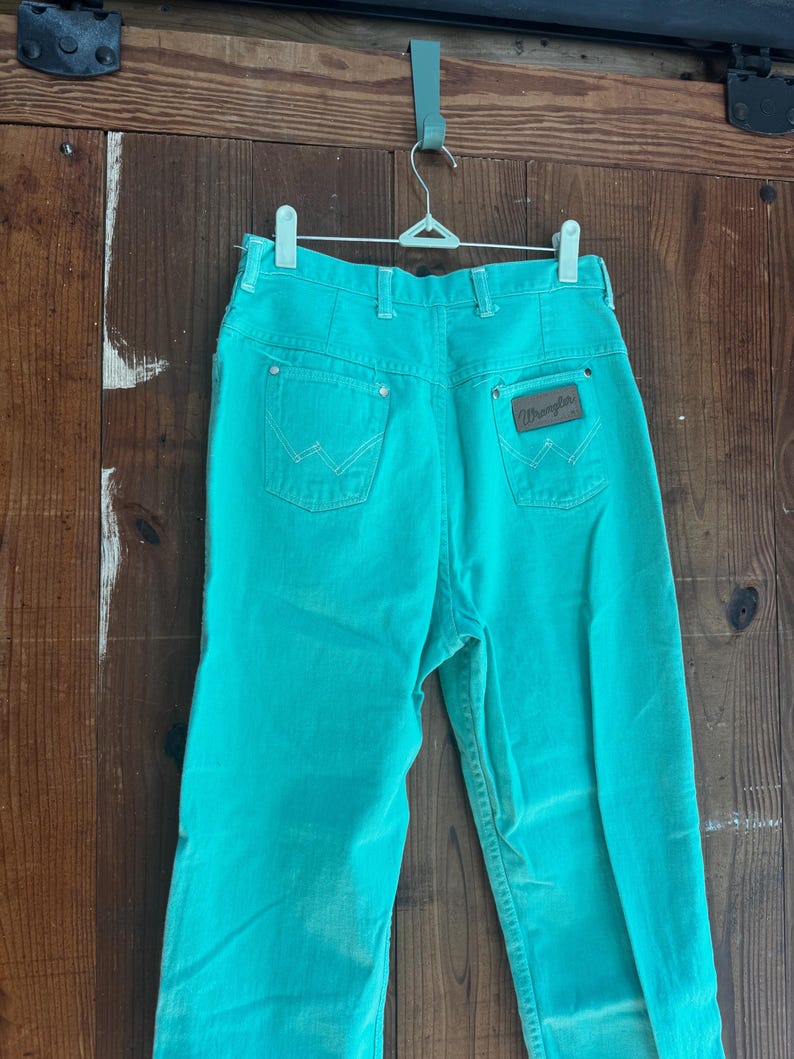 May include: Turquoise Wrangler jeans hanging on a metal hanger. The jeans have two back pockets with the Wrangler logo on the right pocket. The jeans are made of a sturdy material and have a classic fit.