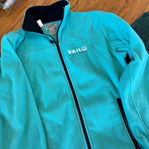 Puede incluir: Una chaqueta de forro polar color turquesa brillante con cremallera frontal negra completa y cuello interior negro. El pecho presenta texto bordado en blanco que dice VAIL COLORADO con un peque&ntilde;o logotipo circular y bolsillos laterales con cremallera.