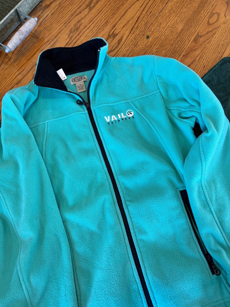 Puede incluir: Una chaqueta de forro polar color turquesa brillante con cremallera frontal negra completa y cuello interior negro. El pecho presenta texto bordado en blanco que dice VAIL COLORADO con un peque&ntilde;o logotipo circular y bolsillos laterales con cremallera.