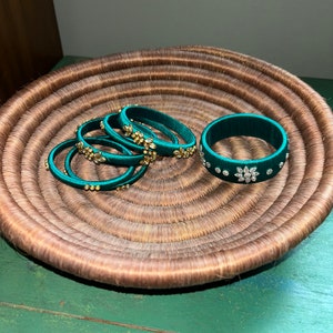 Puede incluir: Un conjunto de seis pulseras de seda verde con adornos dorados y blancos. Las pulseras están apiladas unas sobre otras en una cesta tejida.