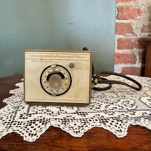 May include: A vintage General Electric Automatic Timer with a white face and brown casing. The timer has a dial with numbers 1 through 12, AM and PM markings, and a knob for setting the time. The timer is on a white lace doily on a wooden table.
