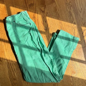 May include: A pair of mint green jeans, laid flat on a wooden floor. The jeans have a classic five-pocket design and a straight leg cut. The fabric appears to be a medium-weight cotton or denim. The sunlight casts shadows across the fabric, highlighting the texture.