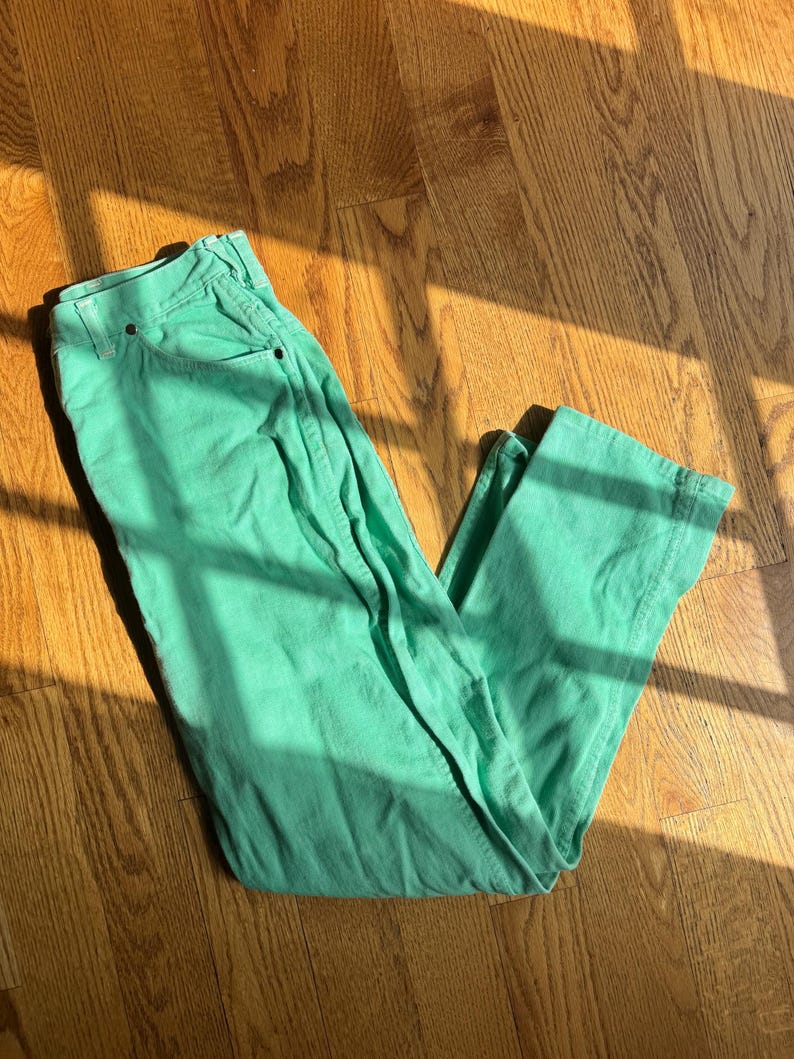 May include: A pair of mint green jeans, laid flat on a wooden floor. The jeans have a classic five-pocket design and a straight leg cut. The fabric appears to be a medium-weight cotton or denim. The sunlight casts shadows across the fabric, highlighting the texture.
