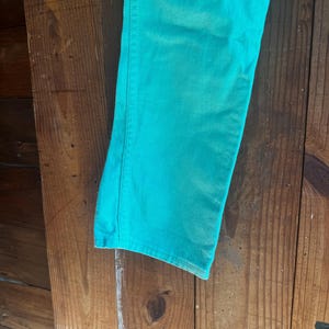 May include: Close-up of a pair of turquoise denim jeans. The jeans are a solid color and appear to be made of a soft denim material. The jeans are hanging against a wooden background.