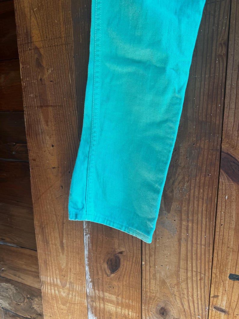 May include: Close-up of a pair of turquoise denim jeans. The jeans are a solid color and appear to be made of a soft denim material. The jeans are hanging against a wooden background.