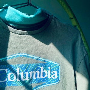 Puede incluir: Una camiseta de manga larga color verde salvia con cuello alto y un parche gr&aacute;fico azul en el pecho. El parche muestra el texto blanco Columbia sobre un fondo azul con l&iacute;neas de contorno topogr&aacute;ficas amarillas, colgada en una percha negra.