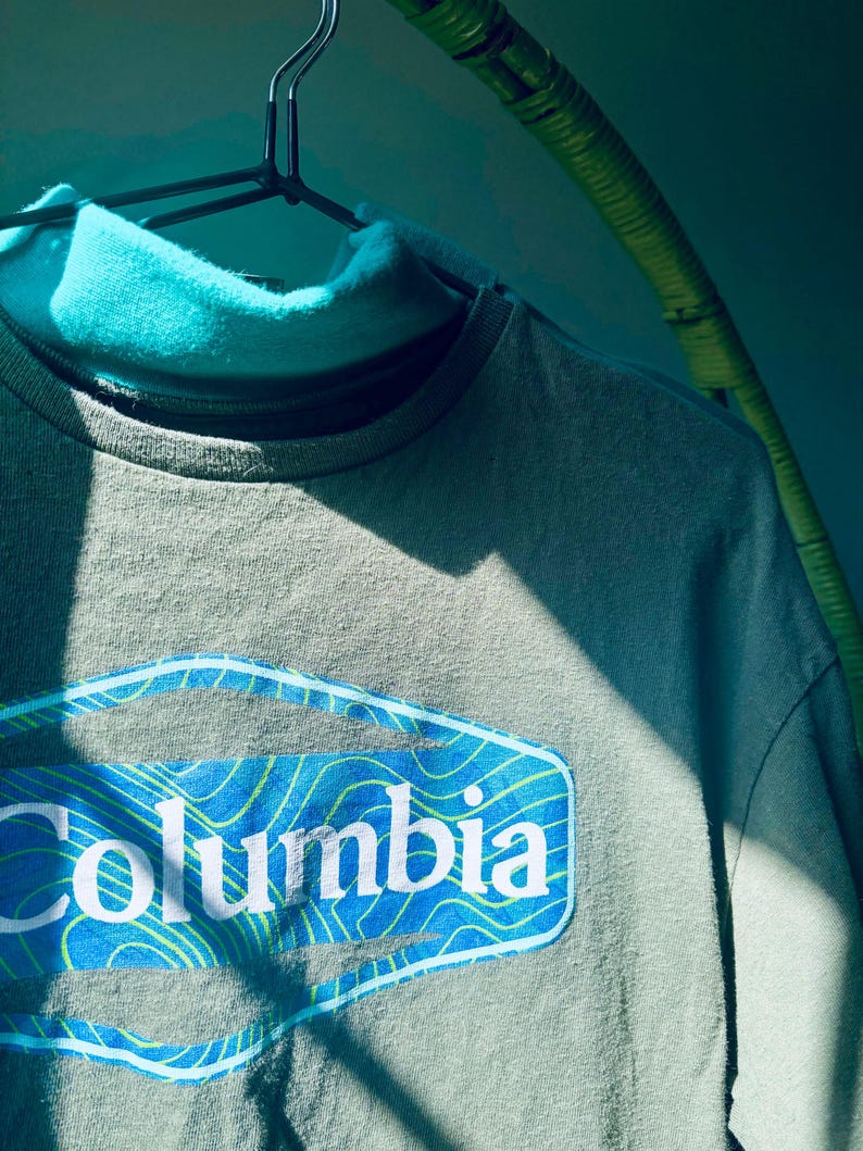 Puede incluir: Una camiseta de manga larga color verde salvia con cuello alto y un parche gr&aacute;fico azul en el pecho. El parche muestra el texto blanco Columbia sobre un fondo azul con l&iacute;neas de contorno topogr&aacute;ficas amarillas, colgada en una percha negra.