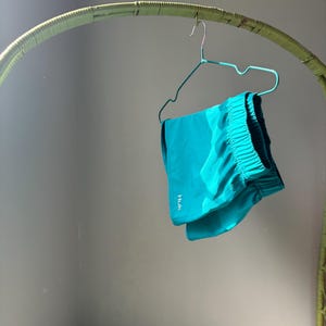 Puede incluir: Unos pantalones cortos deportivos de color turquesa brillante con cintura el&aacute;stica y el texto blanco HUK impreso verticalmente en el lado inferior izquierdo. Los pantalones cuelgan de una percha de alambre azul claro sobre un fondo neutro.
