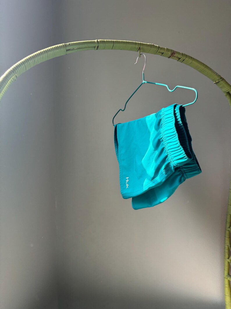 Puede incluir: Unos pantalones cortos deportivos de color turquesa brillante con cintura el&aacute;stica y el texto blanco HUK impreso verticalmente en el lado inferior izquierdo. Los pantalones cuelgan de una percha de alambre azul claro sobre un fondo neutro.