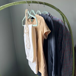 Op de afbeelding: Een groene bamboe kledingrek met vier lichtblauwe hangers met een witte broek, een beige t-shirt, een donkerblauw shirt en een geruit shirt.