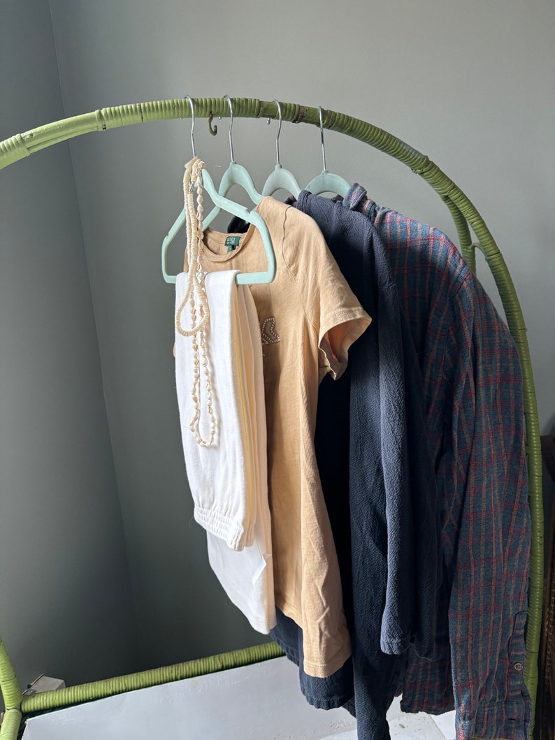 Op de afbeelding: Een groene bamboe kledingrek met vier lichtblauwe hangers met een witte broek, een beige t-shirt, een donkerblauw shirt en een geruit shirt.