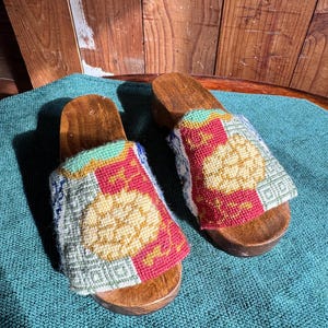 May include: A pair of wooden sandals with a colorful patterned fabric upper. The fabric is a patchwork of different colors and patterns, including red, green, yellow, and blue. The sandals have a wooden sole and a simple design.