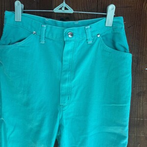 May include: Teal-colored denim pants with a classic five-pocket design. The pants are hung on a white and green hanger against a wooden background. The pants have a button and zipper closure and are made of a sturdy denim material.