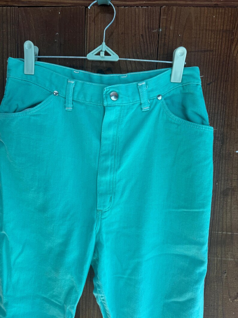 May include: Teal-colored denim pants with a classic five-pocket design. The pants are hung on a white and green hanger against a wooden background. The pants have a button and zipper closure and are made of a sturdy denim material.
