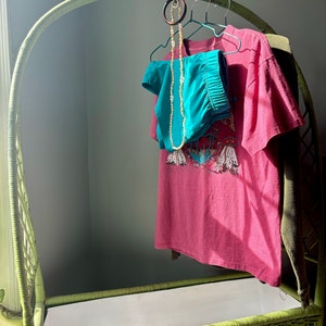 Op de afbeelding: Een groene rieten opbergbank met een witte voering. Een roze T-shirt met een grafische print hangt aan een houten hanger. Een turquoise broek en een kralenketting hangen aan dezelfde hanger.