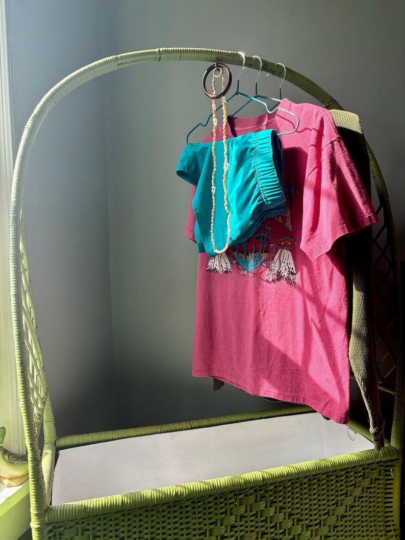 Op de afbeelding: Een groene rieten opbergbank met een witte voering. Een roze T-shirt met een grafische print hangt aan een houten hanger. Een turquoise broek en een kralenketting hangen aan dezelfde hanger.