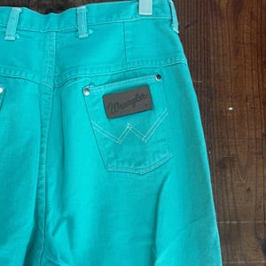 May include: Turquoise Wrangler jeans with a high waist and a brown leather patch on the back pocket. The patch features the Wrangler logo and white stitching. The jeans are hanging on a white hanger.