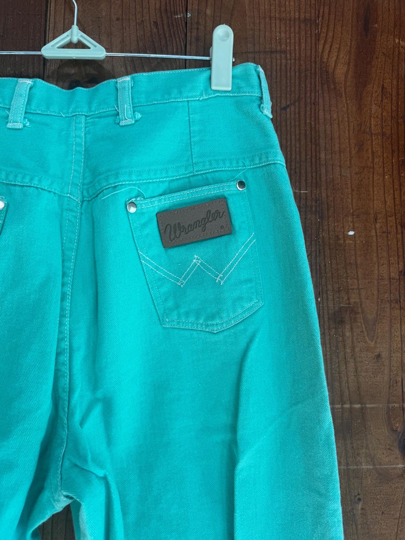May include: Turquoise Wrangler jeans with a high waist and a brown leather patch on the back pocket. The patch features the Wrangler logo and white stitching. The jeans are hanging on a white hanger.
