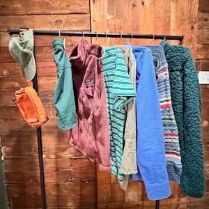 Op de afbeelding: Een zwarte metalen kledingrek met verschillende kledingstukken die eraan hangen. De kleding omvat een ​​groen shirt, een donkerrode fleecejack, een groen-wit gestreept shirt, een lichtblauw shirt, een grijs shirt, een blauw-wit gestreept shirt en een groen fleecejack. Er hangt ook een groene hoed en een oranje heuptas aan de rek.