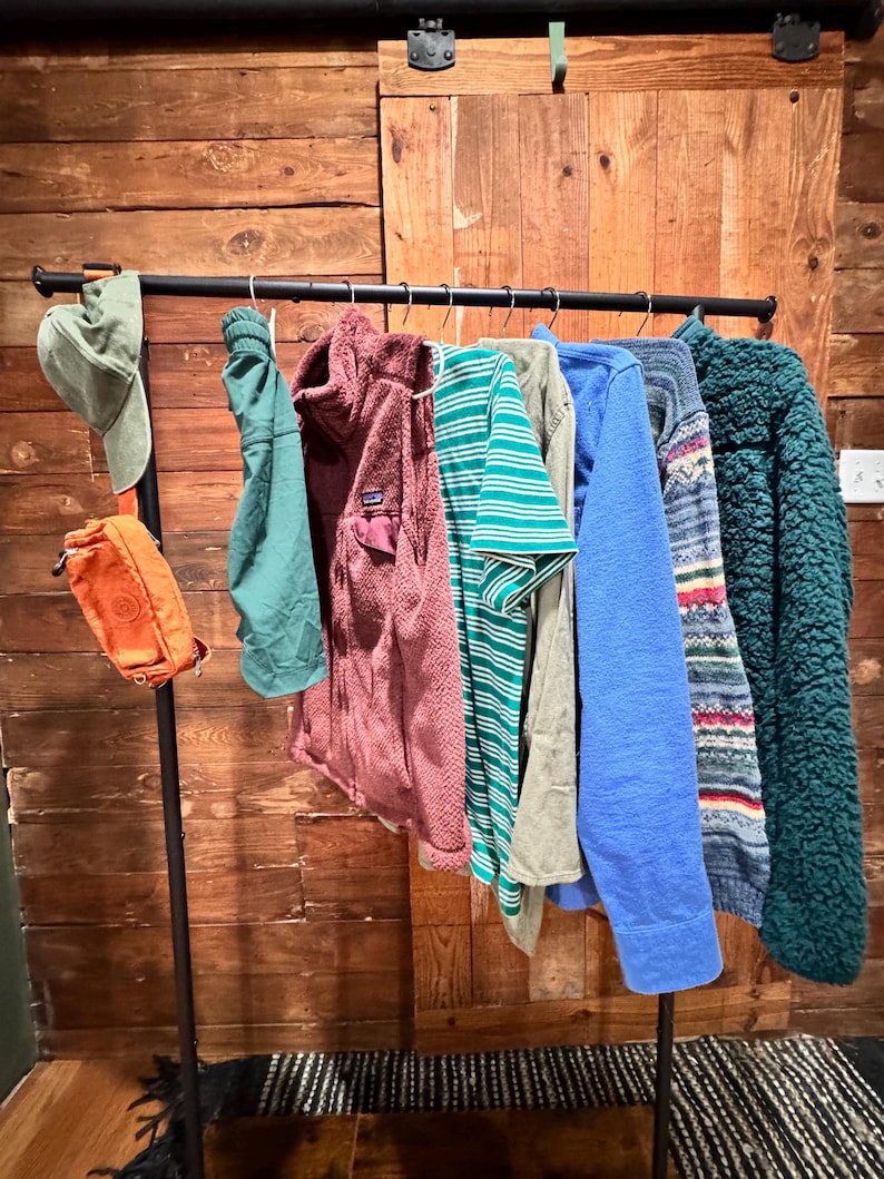 Op de afbeelding: Een zwarte metalen kledingrek met verschillende kledingstukken die eraan hangen. De kleding omvat een ​​groen shirt, een donkerrode fleecejack, een groen-wit gestreept shirt, een lichtblauw shirt, een grijs shirt, een blauw-wit gestreept shirt en een groen fleecejack. Er hangt ook een groene hoed en een oranje heuptas aan de rek.