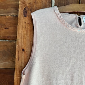 May include: A sleeveless, pale pink knit top with a round neckline. The neckline is embellished with a delicate, beaded trim. The top is hanging on a wooden hanger against a wooden background.