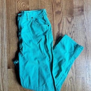May include: A pair of turquoise pants with a button and zipper closure, and front and back pockets. The pants are made of a solid-colored fabric and are folded on a wooden surface.