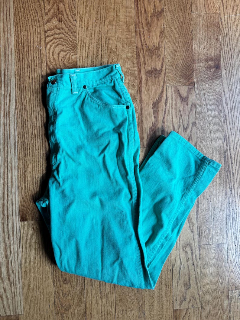 May include: A pair of turquoise pants with a button and zipper closure, and front and back pockets. The pants are made of a solid-colored fabric and are folded on a wooden surface.