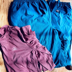 Puede incluir: Una camiseta de manga larga color malva con el logotipo de L.L.Bean en el pecho y un pantal&oacute;n deportivo color verde azulado con cintura el&aacute;stica y un peque&ntilde;o parche rectangular con logotipo en la pierna. Las prendas est&aacute;n sobre madera.