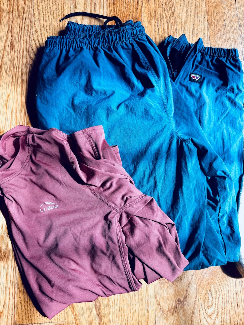 Puede incluir: Una camiseta de manga larga color malva con el logotipo de L.L.Bean en el pecho y un pantal&oacute;n deportivo color verde azulado con cintura el&aacute;stica y un peque&ntilde;o parche rectangular con logotipo en la pierna. Las prendas est&aacute;n sobre madera.