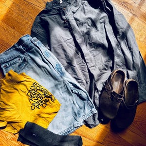 Puede incluir: Una camisa de manga larga gris carb&oacute;n con cuello mao, pantalones vaqueros azul claro, una camiseta amarilla mostaza con el texto negro CALVIN PERFORMANCE, y un par de zuecos de ante marr&oacute;n oscuro con un par de calcetines gris oscuro.