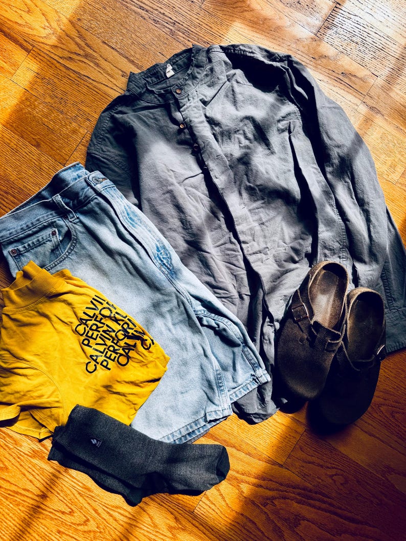 Puede incluir: Una camisa de manga larga gris carb&oacute;n con cuello mao, pantalones vaqueros azul claro, una camiseta amarilla mostaza con el texto negro CALVIN PERFORMANCE, y un par de zuecos de ante marr&oacute;n oscuro con un par de calcetines gris oscuro.
