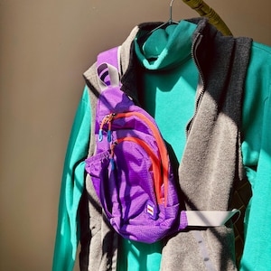 Puede incluir: Una mochila bandolera de color morado brillante con cremalleras naranjas y tiradores azules, sobre un su&eacute;ter de cuello alto color verde azulado y un chaleco de forro polar gris carb&oacute;n. La mochila tiene un parche rectangular peque&ntilde;o.