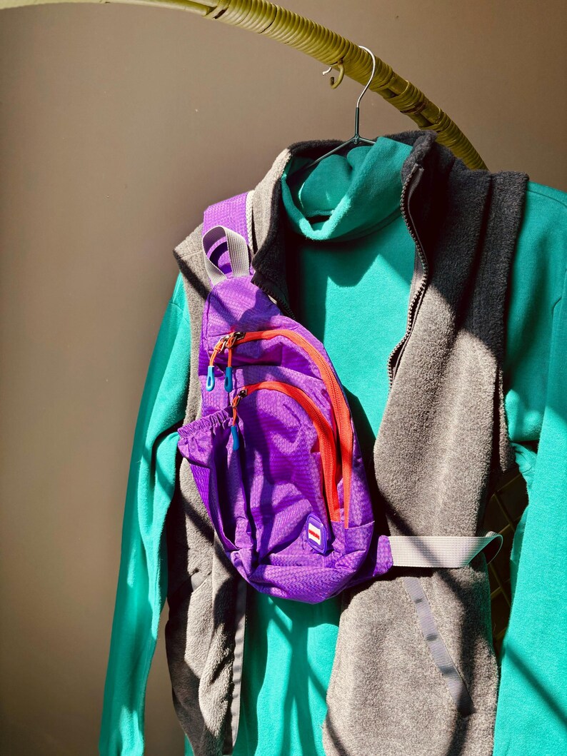 Puede incluir: Una mochila bandolera de color morado brillante con cremalleras naranjas y tiradores azules, sobre un su&eacute;ter de cuello alto color verde azulado y un chaleco de forro polar gris carb&oacute;n. La mochila tiene un parche rectangular peque&ntilde;o.