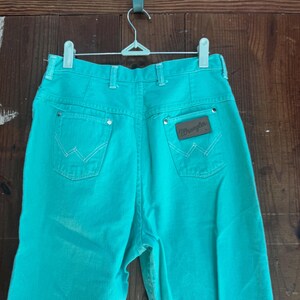 May include: Turquoise Wrangler jeans hanging on a white hanger. The jeans have a high waist, back pockets with white stitching, and a brown leather patch with the Wrangler logo. The jeans are made of a sturdy denim material.