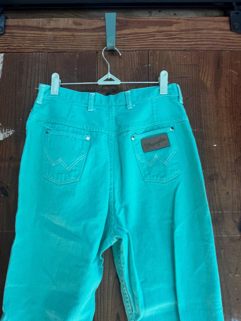 May include: Turquoise Wrangler jeans hanging on a white hanger. The jeans have a high waist, back pockets with white stitching, and a brown leather patch with the Wrangler logo. The jeans are made of a sturdy denim material.