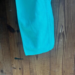 May include: Close-up of a section of turquoise denim jeans. The fabric is a solid, vibrant color with visible texture and stitching along the seam. The jeans are displayed against a rustic wooden background.