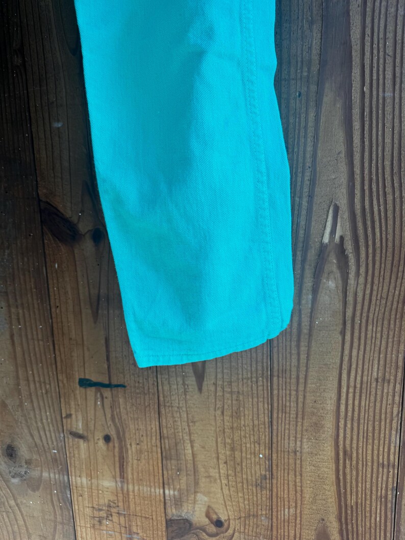 May include: Close-up of a section of turquoise denim jeans. The fabric is a solid, vibrant color with visible texture and stitching along the seam. The jeans are displayed against a rustic wooden background.