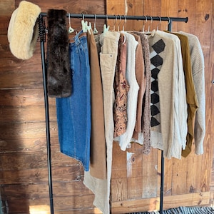 May include: A clothing rack displays a variety of apparel. Items include a faux fur headband, a brown fur scarf, denim jeans, corduroy pants, patterned vest, sweaters, and a long-sleeve shirt. The rack is black, and the background is a wooden door.