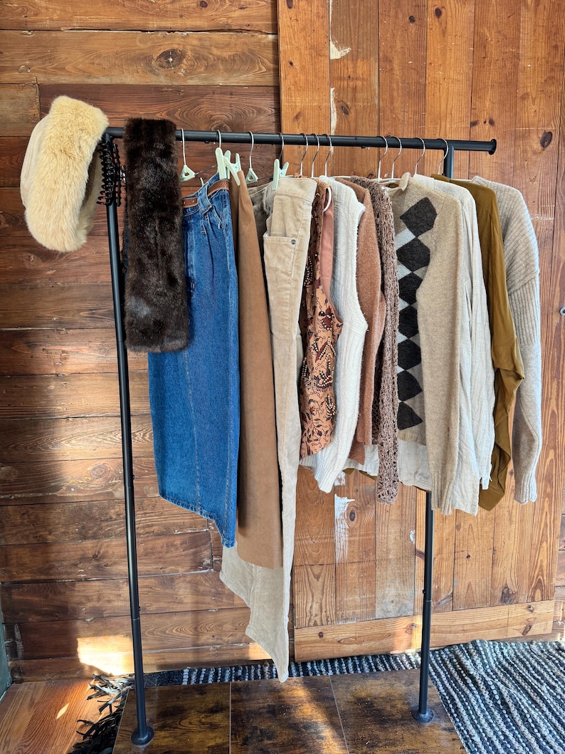 May include: A clothing rack displays a variety of apparel. Items include a faux fur headband, a brown fur scarf, denim jeans, corduroy pants, patterned vest, sweaters, and a long-sleeve shirt. The rack is black, and the background is a wooden door.