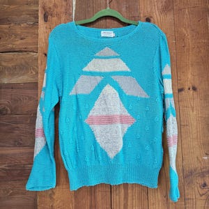 May include: A turquoise knit sweater with a geometric design in white, pink, and gray. The long-sleeved sweater is hanging on a green hanger against a wooden background. The sweater has a round neckline and a textured knit.