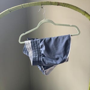 Puede incluir: Unos pantalones cortos deportivos de color azul pizarra con una cintura el&aacute;stica ancha y fruncida, y un ribete blanco en contraste a lo largo del dobladillo curvo. Los pantalones est&aacute;n hechos de una tela ligera y lisa, colgados en una percha verde.