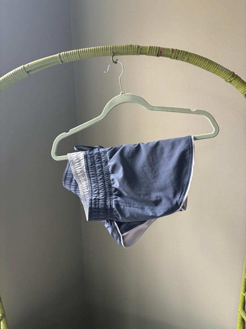 Puede incluir: Unos pantalones cortos deportivos de color azul pizarra con una cintura el&aacute;stica ancha y fruncida, y un ribete blanco en contraste a lo largo del dobladillo curvo. Los pantalones est&aacute;n hechos de una tela ligera y lisa, colgados en una percha verde.
