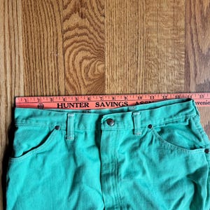 May include: Close-up of a pair of turquoise denim shorts. The shorts have a button and zipper closure, front pockets, and belt loops. A ruler is placed across the top of the shorts, with the text "HUNTER SAVINGS ASSN" visible.