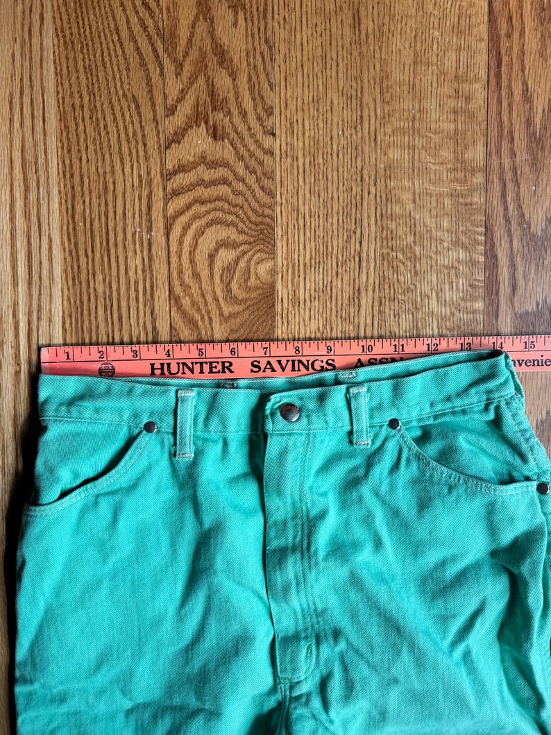 May include: Close-up of a pair of turquoise denim shorts. The shorts have a button and zipper closure, front pockets, and belt loops. A ruler is placed across the top of the shorts, with the text "HUNTER SAVINGS ASSN" visible.