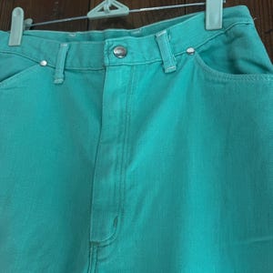 May include: Close-up of turquoise denim jeans. The jeans have a button closure, belt loops, and pockets. The jeans are hanging on a white and gray hanger against a brown wooden background. The jeans are a solid color.